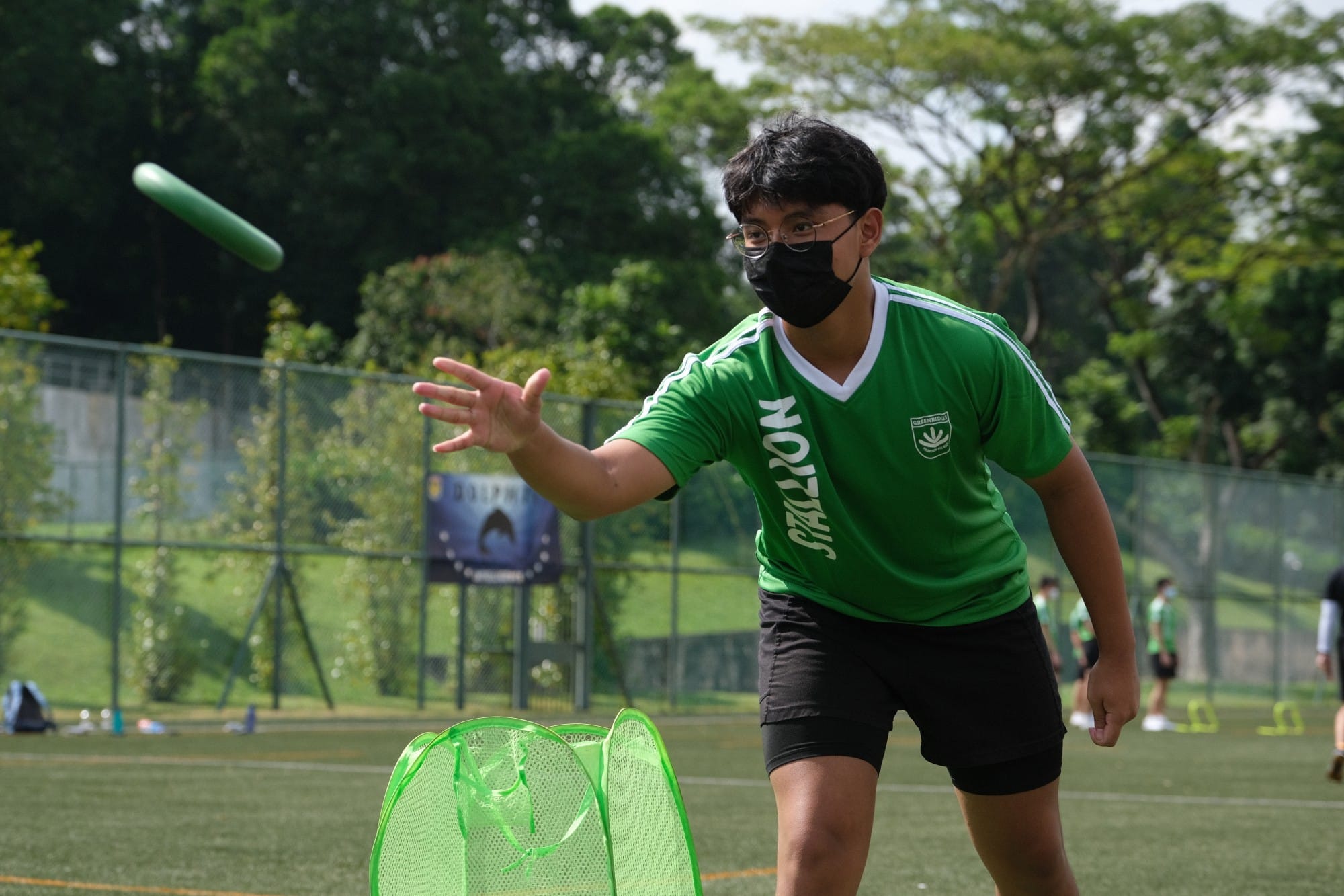 A masked young man in a green jersey throws a green baton toward a green mesh basket on an outdoor field.